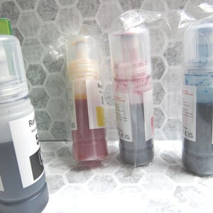 Op de afbeelding: Vier navulflessen met inkt in een transparante plastic verpakking. De flessen bevatten zwarte, gele, magenta en cyaan inkt. Elke fles heeft een wit etiket met zwarte tekst. Op een wit oppervlak met een zeshoekig patroon.