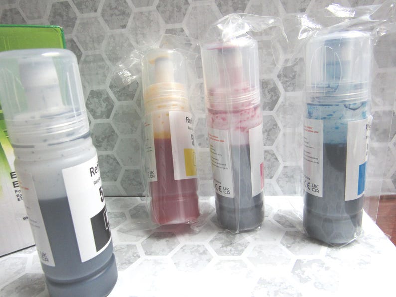 Op de afbeelding: Vier navulflessen met inkt in een transparante plastic verpakking. De flessen bevatten zwarte, gele, magenta en cyaan inkt. Elke fles heeft een wit etiket met zwarte tekst. Op een wit oppervlak met een zeshoekig patroon.