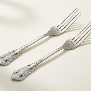 May include: Two silver forks with decorative handles and tines. One fork is engraved with "Mr." and the other with "Mrs." The date "07.22.2024" is engraved on each handle. These forks are likely wedding or anniversary gifts.