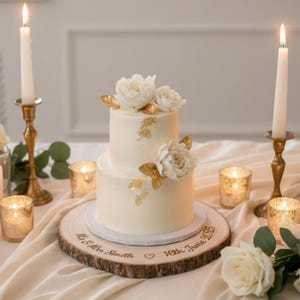 Op de afbeelding: Een witte bruidstaart met twee lagen, versierd met witte bloemen en gouden accenten, op een houten taartstandaard. De standaard is gegraveerd met de namen "Mr & Mrs Smith" en de datum "16th June 2025". Kaarsen en bloemstukken omringen de taart.