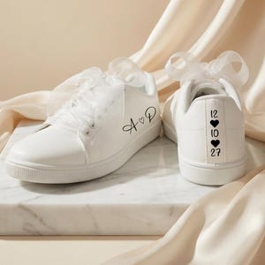 Peut inclure: Une paire de baskets blanches avec des lacets en ruban transparent. Le côté d'une chaussure présente le texte noir A cœur D. Le talon de l'autre chaussure affiche le texte noir 12, un cœur, 10, un cœur et 27 empilés verticalement.