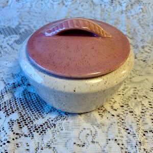 May include: A vintage ceramic lidded bowl with a speckled off-white base and a pink lid. The lid has a handle and a speckled pattern. The bowl is sitting on a white lace tablecloth.