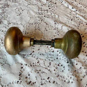May include: A vintage brass door knob set featuring two rounded, weathered brass knobs connected by a central square metal spindle. The hardware displays a tarnished, aged patina with visible surface wear and a classic spherical shape.
