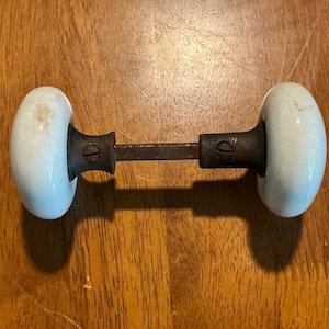 May include: A vintage door knob set featuring two smooth, round, white porcelain knobs connected by a central square metal spindle. Each knob is attached to a dark, weathered metal base with a visible screw, showing signs of age and wear.