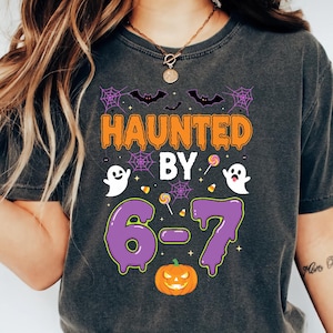 May include: Dark gray t-shirt with a Halloween-themed graphic. The design features the text "HAUNTED BY 6-7" in orange and purple, along with bats, spiderwebs, ghosts, and a jack-o'-lantern. The shirt is a casual wear item.