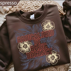 May include: Espresso-colored sweatshirt with the text "RADICALIZED BY BASIC DECENCY" in red and white, surrounded by blue leaves and cream-colored flowers. The sweatshirt has a crew neck and a tag.
