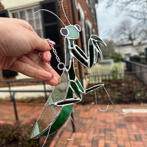 May include: A stained glass praying mantis suncatcher with green and clear glass panels, and silver wire detailing. The insect has a chain for hanging. The background shows a brick walkway and a building.
