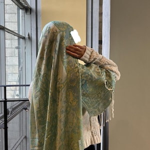 Può includere: Uno scialle a fantasia paisley nei toni del verde, giallo e blu. Lo scialle ha un bordo con frange ed è drappeggiato su una persona che indossa un maglione color crema. Lo sfondo comprende una finestra e un muro.
