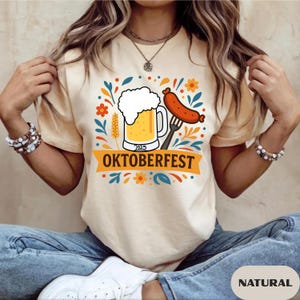 Peut inclure: T-shirt beige clair avec un motif Oktoberfest coloré. Le graphisme comprend une chope de bière avec de la mousse, une saucisse sur une fourchette et le mot "OKTOBERFEST" sur une bannière. Le mot "NATURAL" est imprimé en bas.
