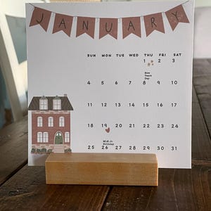 May include: A January calendar with a brown banner that reads "JANUARY". The calendar features a house illustration and is supported by a wooden block. The calendar displays the days of the month, including New Year's Day and MLK Jr. Birthday.