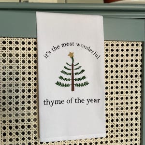 May include: A white rectangular kitchen towel featuring a green embroidered pine tree with a brown trunk and a gold star on top. The text "it's the most wonderful" is arched above the tree, and "thyme of the year" is printed below in black.