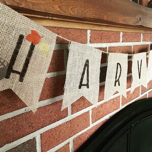 May include: A burlap banner with the word "HARVEST" printed in black letters. The banner is strung across a red brick wall, with a wooden beam above. The letter "H" has orange and yellow leaf accents.