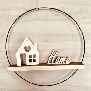 Könnte beinhalten: Ein schwarzer Metallring mit einem daran befestigten Holzregal. Das Regal hat ein weißes Holzhaus mit einem Herz-Ausschnitt und das Wort "Home" in weißen Buchstaben.