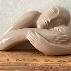 May include: A beige ceramic sculpture of a person in a fetal position, with arms wrapped around the body. The sculpture has smooth, rounded forms and is placed on a wooden surface.
