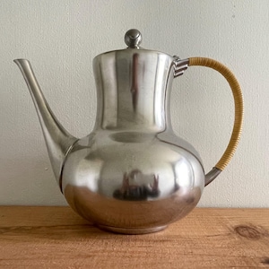 May include: A silver-toned metal coffee pot with a curved spout and a woven handle. The pot has a bulbous body and a lid with a small knob. The handle is made of a light brown woven material. The pot is sitting on a wooden surface.