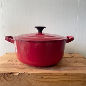 May include: A round, cherry red enameled cast iron Dutch oven with a matching lid and a black knob handle. The pot features two side handles and a smooth, glossy finish, showcasing a classic design for stovetop or oven cooking applications.