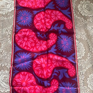 May include: A rectangular textile with a repeating pattern of stylized floral and paisley shapes. The design features vibrant pink and purple elements on a deep burgundy background, with a white border. The textile is likely made of a lightweight fabric.