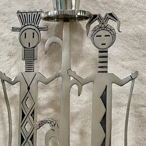 May include: A silver-toned metal candle holder featuring two stylized human figures with outstretched arms. The figures have geometric designs and are connected by a central stem holding a candle cup. The design has a Native American aesthetic.