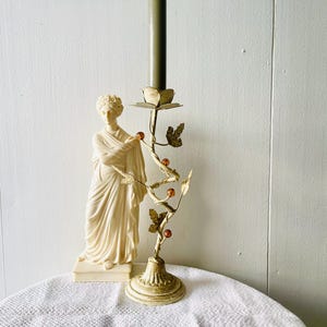 May include: A cream-colored figurine of a person in draped robes standing next to a gold-toned metal candle holder. The candle holder features a vine-like stem with metal leaves and small amber-colored spherical accents, topped with a candle.