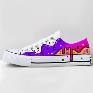 May include: White canvas low-top sneaker with a colorful, artistic design. The design features a purple, orange, and pink dripping pattern on the side, with black dots and stars. The laces are white.