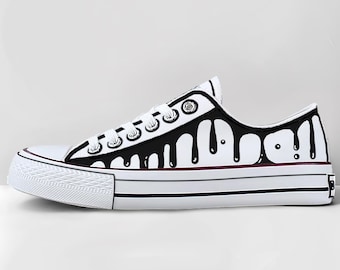 May include: White canvas sneaker with black dripping paint design. The low-top shoe features white laces, a white rubber sole, and a black and burgundy stripe. The design includes the word "Drip" in black.