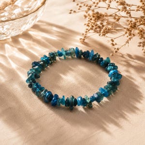 Op de afbeelding: Een cirkelvormige armband bestaande uit talloze kleine, onregelmatige, gepolijste blauwe apatiet edelsteenchips die aan elkaar zijn geregen. De stenen vertonen variërende tinten diep oceaanblauw, taling en doorschijnend cyaan met een getextureerd uiterlijk.