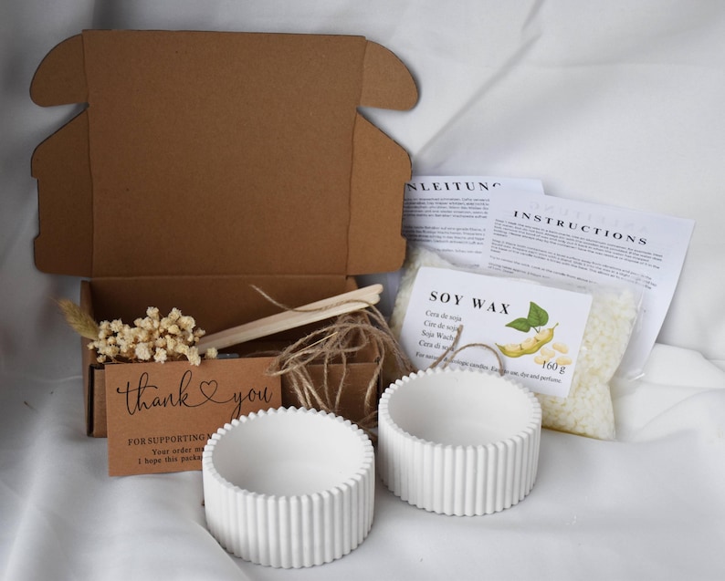 K&ouml;nnte beinhalten: Ein brauner Karton mit einer Dankeskarte, zwei wei&szlig;en Beton-Kerzenhaltern, einem Beutel mit Sojawachs-Flocken und Anleitungen zum Kerzenmachen. Der Sojawachsbeutel tr&auml;gt den Text "Soy Wax" und "160g".
