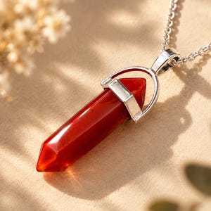 Op de afbeelding: Een ketting met hanger bestaande uit een gepolijste, dubbelzijdige, dieprode carneool kristallen punt. De steen is bevestigd in een zilverkleurige metalen kap en hangt aan een bijpassende zilverkleurige metalen schakelketting.