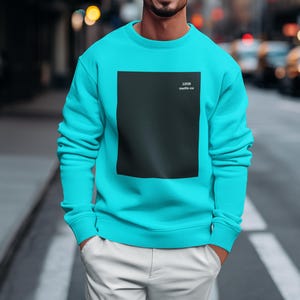 Puede incluir: Una sudadera turquesa con un gran cuadrado negro en la parte delantera. El texto "1206 matte-co" está impreso en blanco dentro del cuadrado negro. La persona lleva pantalones blancos y tiene las manos en los bolsillos.