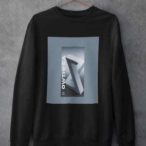 Puede incluir: Sudadera negra con un diseño gráfico rectangular. El diseño presenta un diseño geométrico en escala de grises con el texto "OWTHI-CA" en blanco. La sudadera está colgada en una percha de color cobre contra una pared gris.