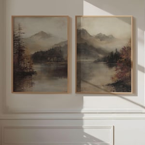 May include: Two framed landscape paintings. Each depicts a tranquil lake scene with mountains in the background. The artwork uses a muted colour palette of browns, greens, and greys, creating a vintage aesthetic. The frames are a light wood colour.