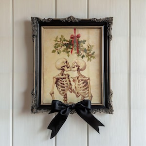 Könnte beinhalten: Gerahmter Druck mit zwei Skeletten, die sich unter einem Stechpalmenzweig mit roter Schleife küssen. Der schwarze, verzierte Rahmen hat eine schwarze Satinschleife. Das Kunstwerk hat eine Vintage-Gothic-Ästhetik.