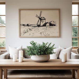 May include: A framed black and white sketch of a windmill in a field hangs above a light-colored sofa. A wooden coffee table displays a large, round, light-colored planter with greenery, and three white candles. Two wicker chairs sit on either side of the table.