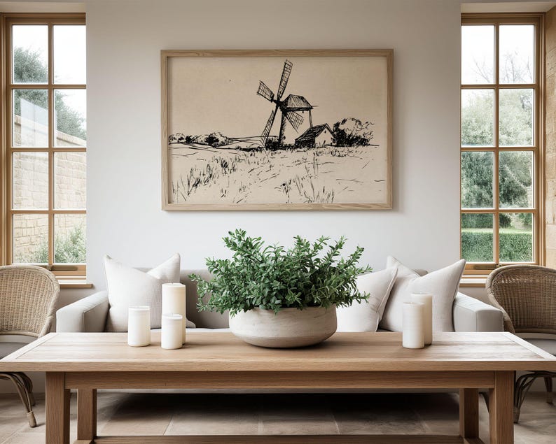 May include: A framed black and white sketch of a windmill in a field hangs above a light-colored sofa. A wooden coffee table displays a large, round, light-colored planter with greenery, and three white candles. Two wicker chairs sit on either side of the table.