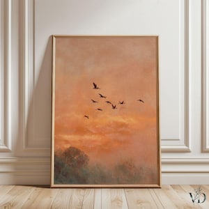 Peut inclure: Une impression d'art encadrée représentant un coucher de soleil avec des oiseaux en vol. L'œuvre d'art présente une palette de couleurs chaudes d'orange, de pêche et de marron. Les oiseaux se détachent sur le ciel, et l'impression est exposée sur un sol en bois.