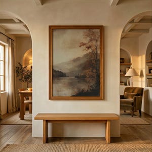 May include: A rectangular wooden framed wall art print depicting a misty landscape with a calm lake, rolling mountains, and autumn-toned trees. The artwork is mounted on a smooth, light-colored wall above a simple, solid light oak wooden bench.