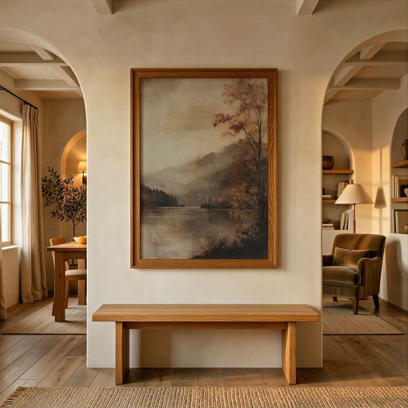 May include: A rectangular wooden framed wall art print depicting a misty landscape with a calm lake, rolling mountains, and autumn-toned trees. The artwork is mounted on a smooth, light-colored wall above a simple, solid light oak wooden bench.