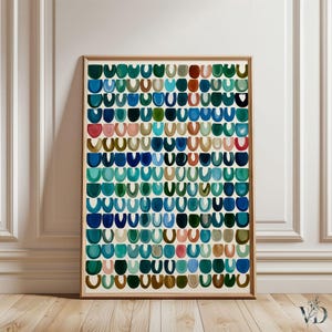 May include: Abstract art print featuring a pattern of colorful, semi-circular shapes in shades of blue, green, pink, and brown. The artwork is framed in a light wood frame and displayed against a white wall and wooden floor.