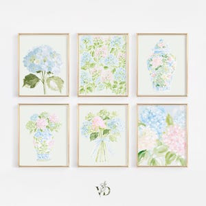 Può includere: Un set di sei stampe artistiche da parete incorniciate con illustrazioni ad acquerello di fiori di ortensia blu e rosa, alcuni disposti in bouquet e altri in vasi di porcellana blu e bianca, il tutto racchiuso in cornici dorate.