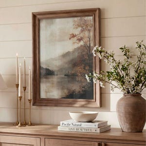 May include: A framed landscape painting depicting a misty lake surrounded by trees with autumn foliage, displayed alongside four brass candlesticks, a white ceramic bowl, and two stacked books titled Pacific Natural and Live Beautiful.
