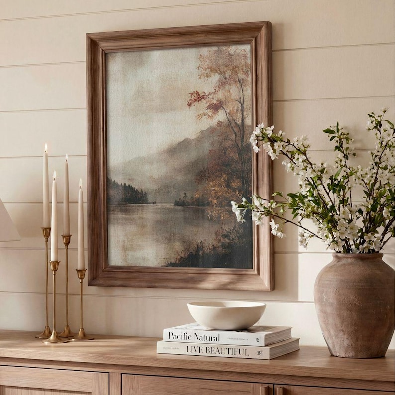 May include: A framed landscape painting depicting a misty lake surrounded by trees with autumn foliage, displayed alongside four brass candlesticks, a white ceramic bowl, and two stacked books titled Pacific Natural and Live Beautiful.