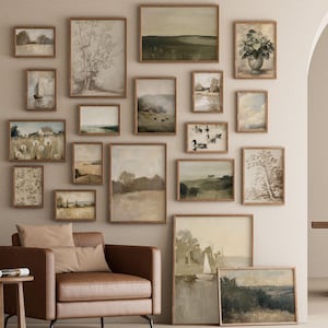 May include: A collection of framed landscape art prints in various sizes and styles, arranged on a neutral-colored wall. The prints feature muted tones of green, brown, and beige, with subjects including trees, fields, and water scenes. A brown leather armchair and side table are in the foreground.
