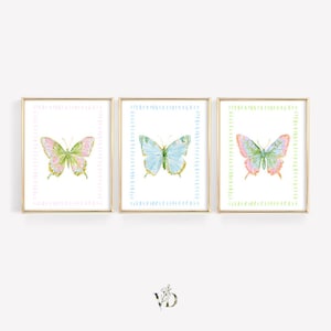 May include: A set of three framed wall art prints, each featuring a watercolor butterfly centered within a dashed rectangular border. The left print has a pink border, the center has a blue border, and the right has a light green border.