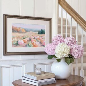 May include: A framed painting of a pumpkin patch in a field, with a gold-colored frame, hangs on a white paneled wall. A white vase holds pink and white hydrangeas. A small glass and stacked books sit on a wooden table.