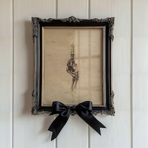 May include: A framed print featuring a skeletal hand holding a lit candle. The artwork is in a black ornate frame with a gold inner border. A black satin bow is tied below the frame, adding a decorative touch. The print is mounted on a white wall.
