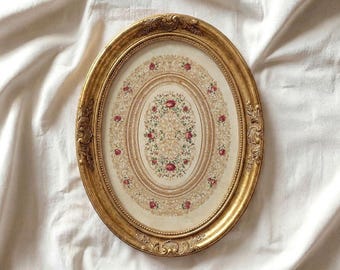Könnte beinhalten: Ein ovaler, goldfarbener Bilderrahmen mit aufwendigen Details. Das gerahmte Kunstwerk zeigt ein Blumendesign mit roten Rosen und grünen Blättern auf cremefarbenem Hintergrund. Der Text "VINTAGE DISCO" und "Curated Timeless Art" steht unten.