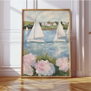 May include: A framed painting depicts a coastal scene with sailboats on blue water. White sailboats with raised sails are in the distance. Pink and white flowers are in the foreground. The painting is in a gold frame and leans against a white wall.