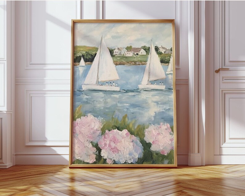 May include: A framed painting depicts a coastal scene with sailboats on blue water. White sailboats with raised sails are in the distance. Pink and white flowers are in the foreground. The painting is in a gold frame and leans against a white wall.
