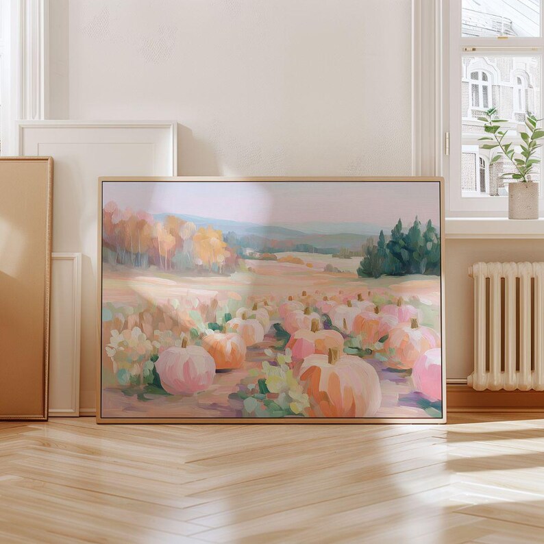 May include: A framed canvas art print depicts a pumpkin patch scene. The artwork features a field of pumpkins in shades of pink and orange, with a backdrop of trees and a soft, pastel color palette. The frame is a light wood color.