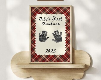 May include: A framed print with a red and white tartan border. The print features the text "Baby's First Christmas" above two handprints and the year "2025". The frame is displayed on a wooden shelf.